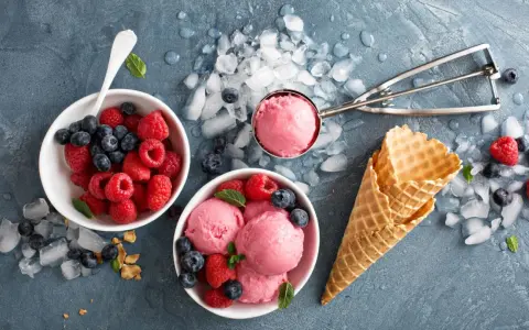 Himbeeren und Blaubeeren neben Schalen mit leckerem Milcheis selber machen. Eiswaffeln und ein Eisportionierer liegen bereit.