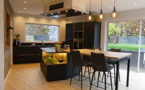 Cuisine noir et bois, mur gris texturé. Îlot central avec éclairage, table haute et tabourets. Fenêtre donnant sur jardin.