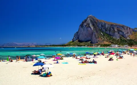 Spiaggia affollata con ombrelloni colorati, gente che si gode le vacanze al mare con bambini, acque turchesi e una montagna imponente sullo sfondo.