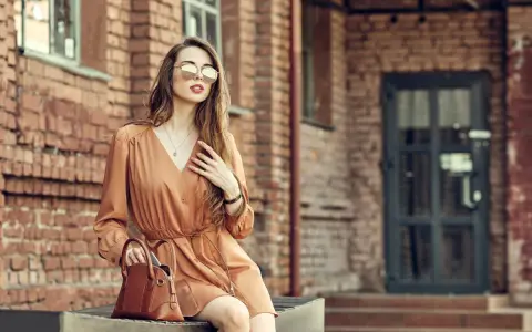 Elegancka kobieta w brązowej sukience i okularach przeciwsłonecznych, z torebką. Idealne buty do brązowej sukienki podkreślają jej styl.