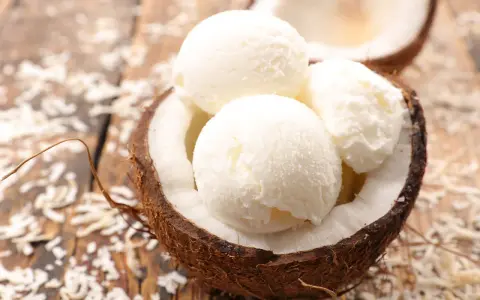 Cremiges Kokosnuss Eis in einer halbierten Kokosnuss, umgeben von Kokosraspeln auf Holz. Ein tropischer Genuss!