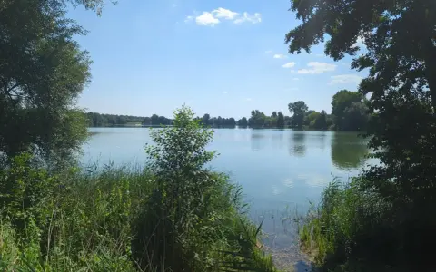 Spokojne jezioro Małe Żnińskie otoczone zielenią, z błękitnym niebem i białymi chmurami odbijającymi się w wodzie.