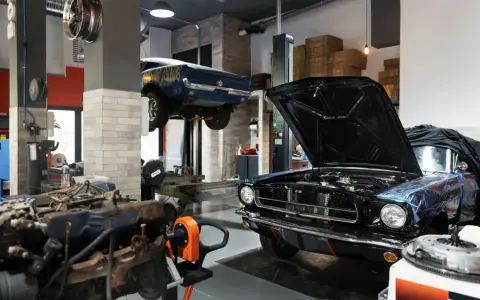 Un taller ordenado con un coche clásico negro con el capó abierto y otro azul en elevación. Motores y herramientas a la vista.