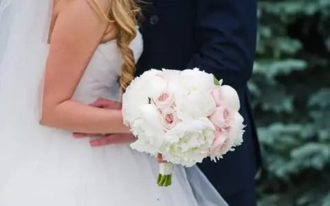 Piękny bukiet ślubny piwonie i eustoma w dłoniach pary młodej. Delikatne białe i różowe kwiaty tworzą romantyczną kompozycję.