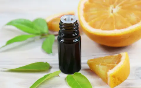 Flacon d'huile essentielle de citrus sinensis, entouré de feuilles vertes et de morceaux d'orange fraîche sur fond de bois clair.