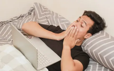 Mężczyzna w łóżku z laptopem, zakrywający usta dłońmi, może symbolizować walkę z uzależnieniem od pornografii.