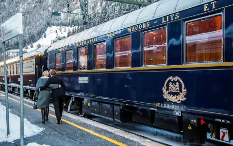 Ett par omfamnar varandra vid en snötäckt station, redo för en resa med Orientexpressen. Tågets mörkblå vagnar med gyllene detaljer vittnar om dess klassiska orientexpressen rutt.