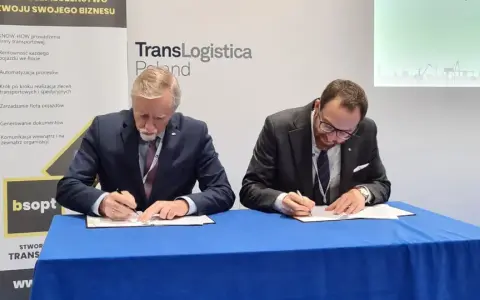 Dwóch mężczyzn w garniturach podpisuje dokumenty. Obok baner z napisem TransLogistica Poland, sugerujący umowę w branży gpw logistics.
