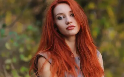 Eine Frau mit leuchtend roten Haaren steht im Freien. Ihre langen, feurigen Haare umrahmen ihr Gesicht, während sie in die Kamera blickt. Die Natur im Hintergrund ist unscharf und zeigt herbstliche Farben.