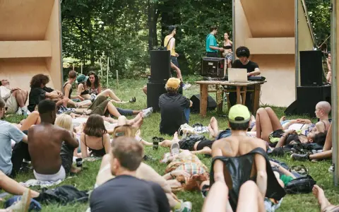 Un gruppo di persone rilassate su un prato in un **boutique festival**, con un DJ che suona musica in sottofondo.