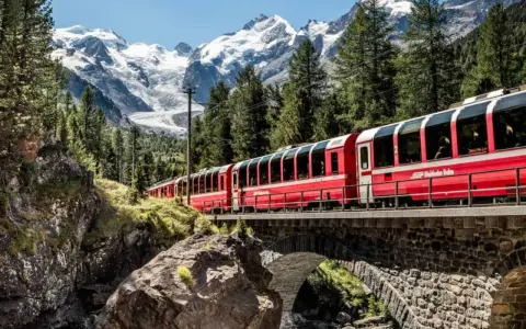 A red train travels across a stone bridge, winding its way from Chur to Tirano through a stunning alpine landscape with snow-capped mountains.