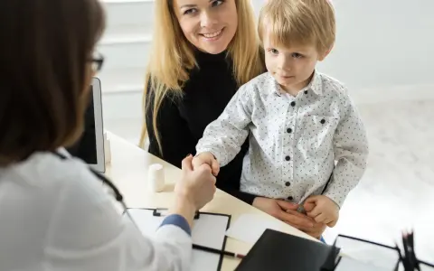 Mama z synkiem u lekarza. Chłopiec ściska dłoń lekarce, a mama uśmiecha się. W tle widać tablet i białą buteleczkę, jakby z lekiem. To ważny moment, być może wpis do książeczki zdrowia.