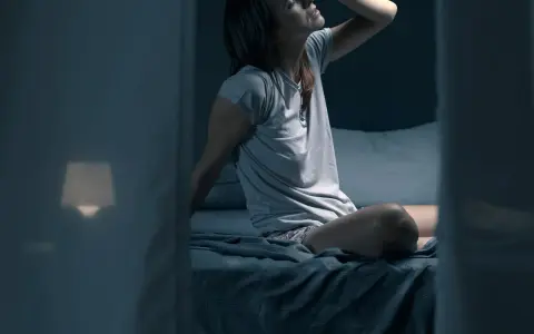 Frau sitzt im Bett, die Stirn gerunzelt, die Hand an der Stirn. Sie fragt sich, wie viel schwitzt man in der Nacht.