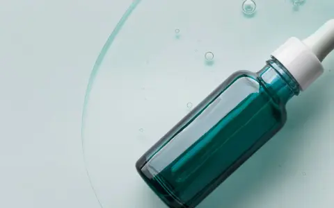 Szklana buteleczka z pipetką, wypełniona zielonym serum, na tle płynu z bąbelkami.