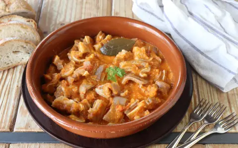 Guiso casero de oreja de cerdo en salsa de tomate, servido en cazuela de barro, acompañado de pan rústico y tenedores.