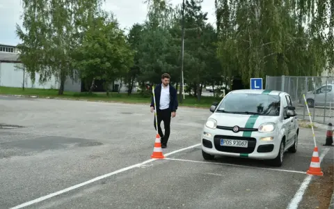 Młody mężczyzna ustawia pachołek na placu manewrowym. Biały Fiat Panda z literą "L" na dachu czeka na swój egzamin praktyczny kat b.