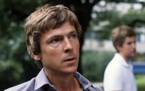 Carl Ivar Nilsson, en man med rufsigt hår och en randig skjorta, tittar bort. En annan man syns suddigt i bakgrunden.