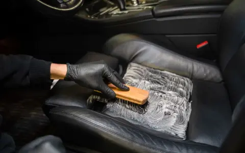 Mano enguantada cepilla asiento de cuero negro con espuma, mostrando cómo limpiar asientos de cuero para un coche impecable.