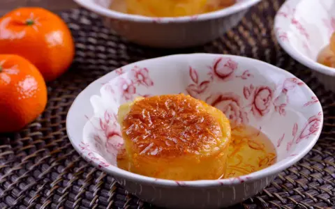 Un esponjoso baba al ron bañado en almíbar, adornado con ralladura de naranja, en un cuenco floral. Cerca, mandarinas frescas.
