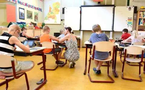 Dzieci siedzą w klasie, ucząc się. Nauczycielka pochyla się nad jednym z uczniów. Ile jest klas w Anglii? Tego nie wiemy, ale ta lekcja wygląda na interesującą.