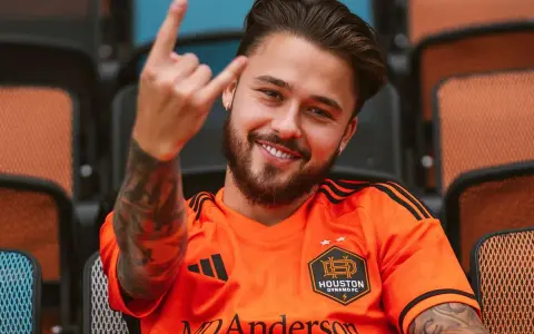 Uśmiechnięty Adam Kowalczyk w koszulce Houston Dynamo FC pozdrawia gestem rock and rolla, siedząc na trybunach.
