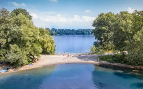 Sommerliche Idylle am Strandbad Wedau: Menschen genießen den Tag am Seeufer, einige radeln, andere entspannen im Sand.