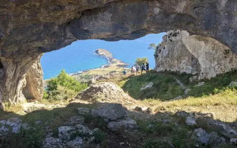 Vista desde el "ojo del diablo", una cueva natural, hacia un paisaje costero con un istmo y el mar azul. Un grupo de personas observa el panorama.