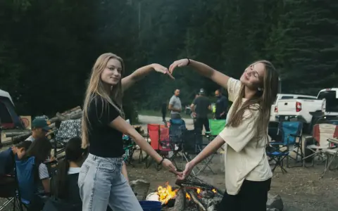 Dwie dziewczyny tworzą serce z dłoni na tle ogniska i lasu. To idealny okres na wakacjach, pełen radości i wspólnych chwil.