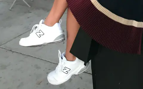 White New Balance dad shoes with a black "N" logo, paired with a pleated skirt.