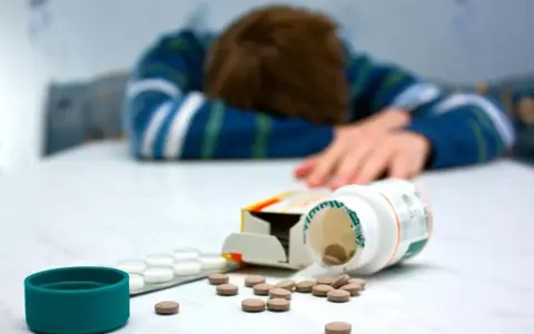 Osoba w bluzie leży na stole, obok rozsypane tabletki, w tym opakowanie leku Seronil 10 mg.