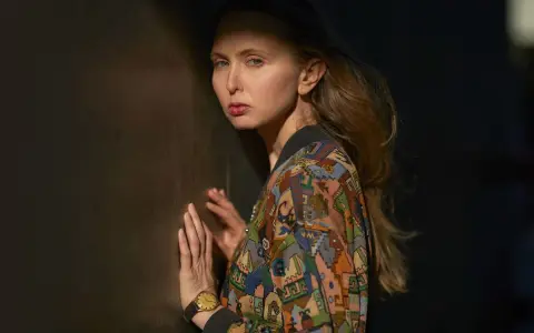 Justyna Wasilewska w kolorowej kurtce opiera się o ciemną ścianę, jej twarz oświetla cień.