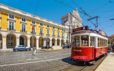 Czerwony tramwaj w Lizbonie, obok żółtego budynku z arkadami. Czy do Portugalii trzeba paszport? Nie, wystarczy dowód osobisty.