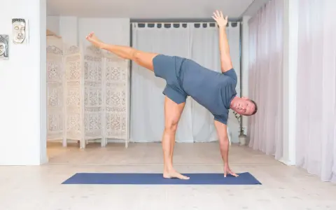 Man utför en dynamisk yoga-position på en blå matta i ett ljust rum med vita draperier och en vikbar rumsavdelare.