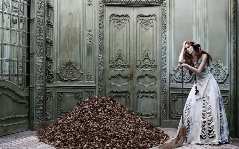 A woman in a tattered gown leans on a broom beside a mountain of leaves in a grand, ornate room, a scene captured by Eugenio Recuenco.