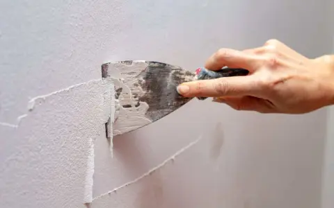 Mano che raschia via pittura lavabile dal muro con una spatola, mostrando il processo di rimozione.