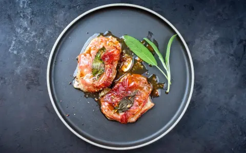 Zwei saftige, im Ofen gebackene italienische Fleischgerichte, umhüllt von knusprigem Schinken und frischem Salbei, serviert auf einem dunklen Teller.