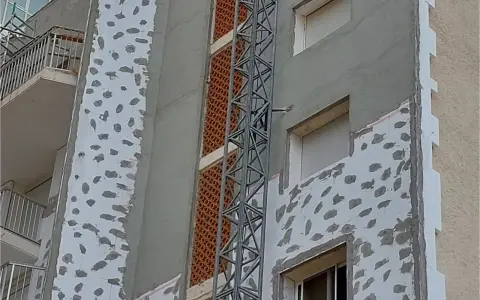Proceso de como aislar una pared ya construida con placas de poliestireno y mortero. Se observa una grúa de obra.