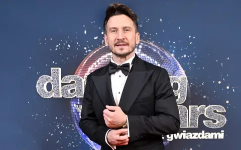 Tomasz Barański w czarnym smokingu pozuje na tle błyszczącej kuli dyskotekowej i napisu "Dancing with the Stars".
