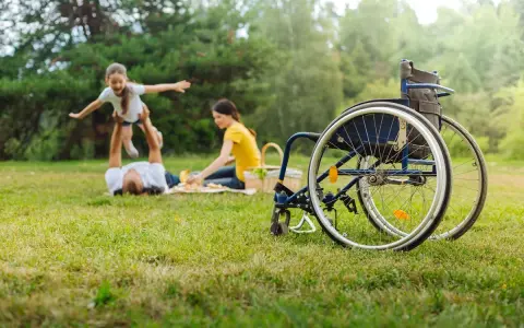 Rodzina na pikniku. Ojciec podrzuca córkę, matka przygotowuje jedzenie. Obok stoi wózek inwalidzki, symbol wsparcia i obecności opiekuna osoby niepełnosprawnej.