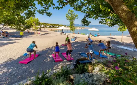 Grupa osób ćwiczy jogę na plaży w Omisalj. W tle widać morze i kemping.