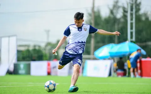 Piłkarz w białej koszulce z niebieskimi rękawami i spodenkami kopie piłkę na zielonym boisku.