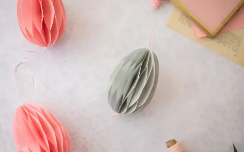Ostereier basteln Papier: Dekorative Papier-Ostereier in Rosa und Grau hängen an Schnüren. Ein Garnknäuel und eine Schere liegen daneben.
