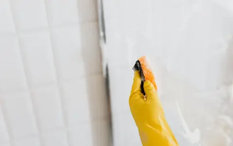 Ręka w żółtej rękawiczce czyści szklaną kabinę prysznicową gąbką. Dowiedz się, czym umyć kabinę prysznicową ze szkła, by lśniła czystością.
