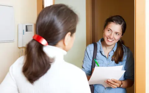 Assistente sociale sorridente, con fogli e penna, parla con una donna alla porta. Il suo lavoro è importante.