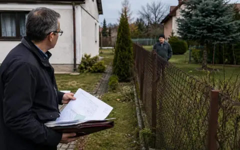 Mężczyzna z dokumentami analizuje teren, szukając sposobu, jak podważyć zasiedzenie. Sąsiad obserwuje go zza płotu.