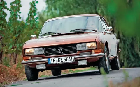 Klasyczny brązowy Peugeot 504 Coupe jedzie po krętej drodze wśród zieleni.