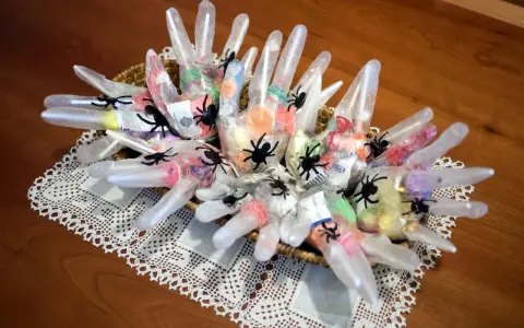 Manualidades chuches Halloween: guantes transparentes rellenos de caramelos y decorados con arañas negras, presentados en una cesta sobre un mantel de encaje.