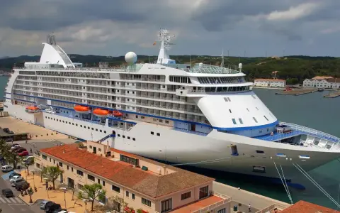 Grande nave da crociera attraccata nel porto Mahon, con edifici storici e colline verdi sullo sfondo.