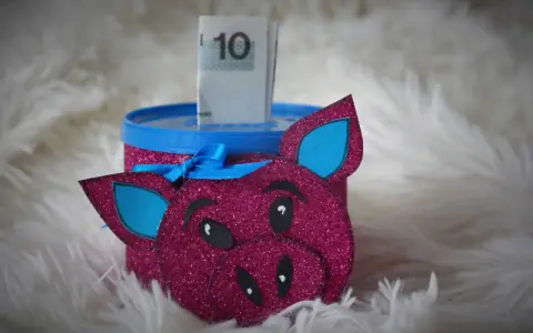Urocza skarbonka diy w kształcie różowej świnki z brokatem, z niebieskimi uszami i kokardką. W środku widać banknot 10 zł.