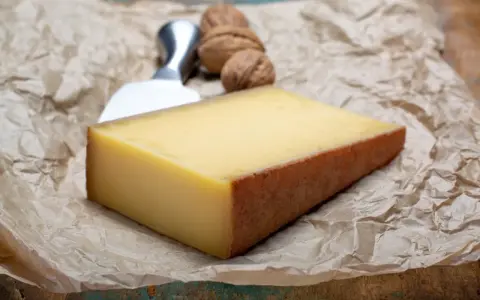 Kawałek żółtego sera z brązową skórką, nóż do sera i orzechy włoskie na papierze.